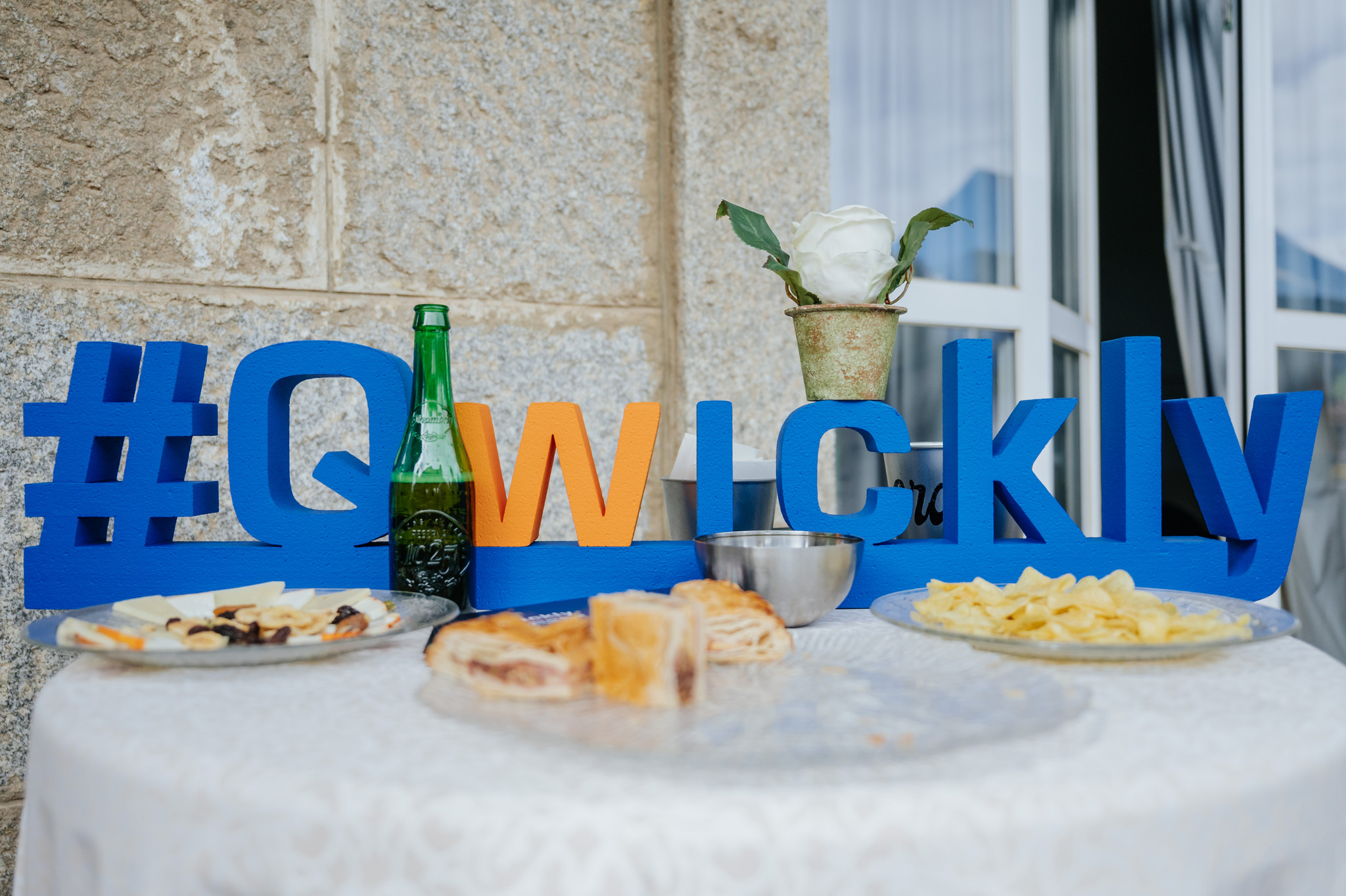 A table in front of a large glass window set with hors d'oeuvres, a glass bottle, and a decorative potted flower, featuring a large blue hashtag sign that reads Qwickly.